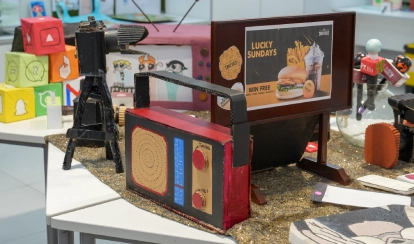 A display table features colourful student-made models, including a cardboard radio, camera, and advertisement board, surrounded by posters and craft cubes decorated with social media icons